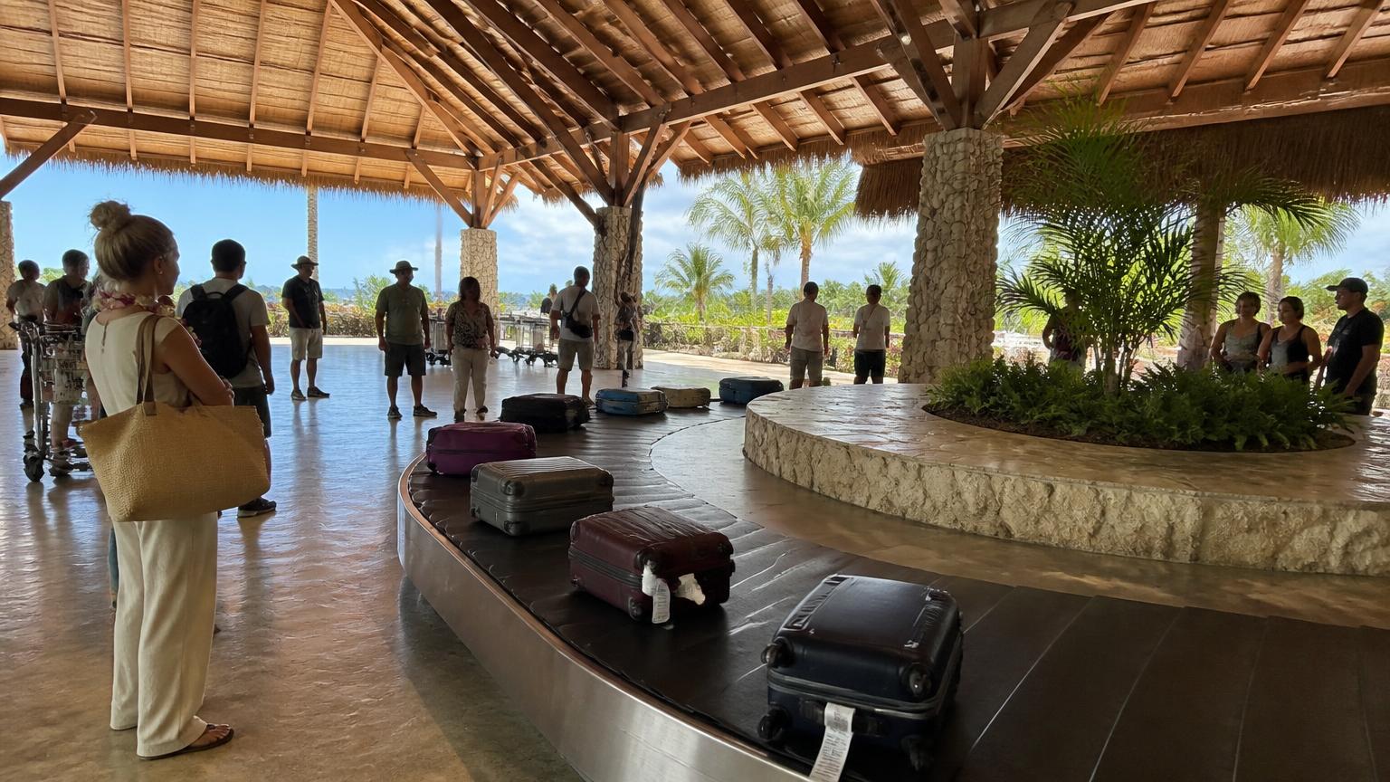 Baggage carousel under a thatched roof at a tropical Caribbean airport with travellers waiting