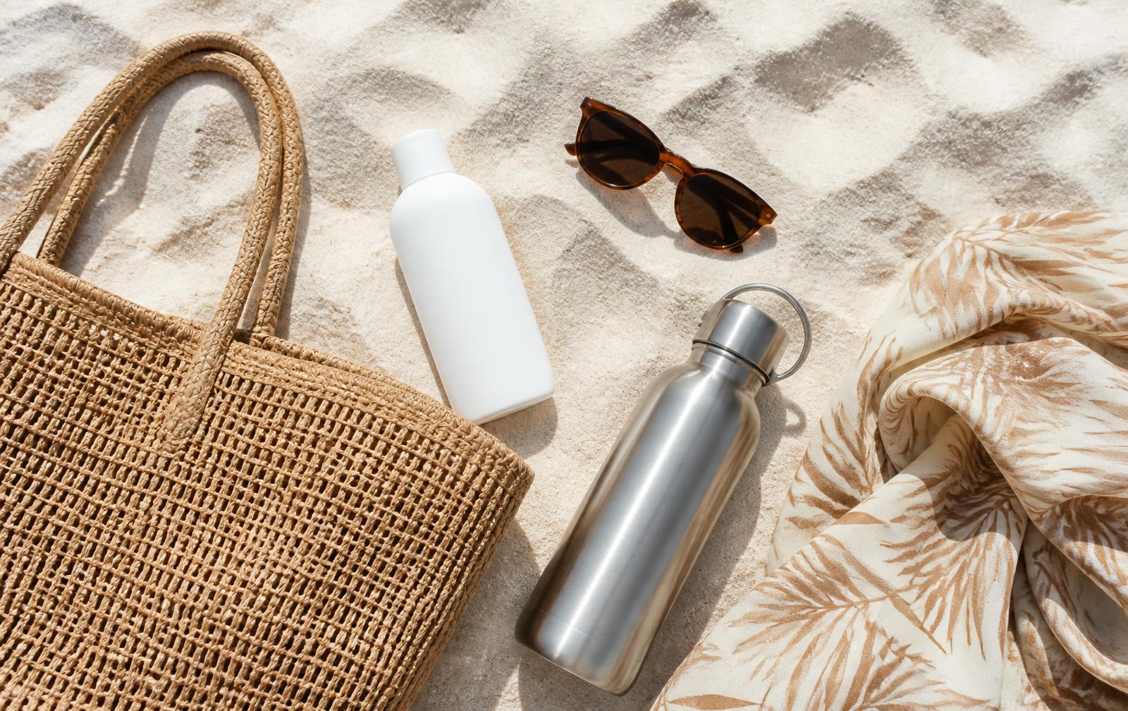 Beach essentials on white sand: tote, sunscreen, sunglasses, water bottle